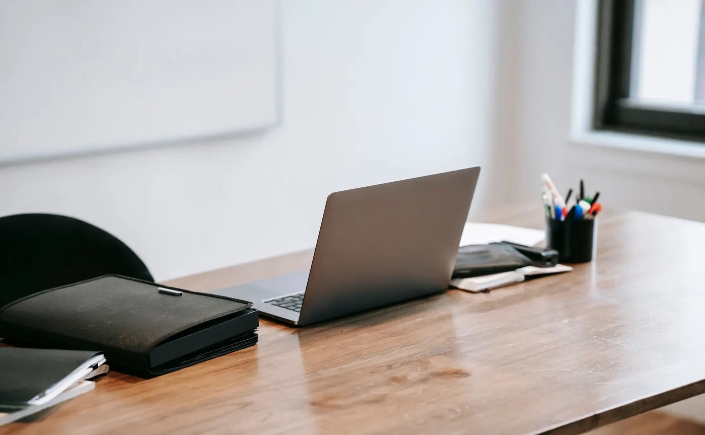 Clean wooden office table with an open laptop, portfolio, notebooks, and cup of pens