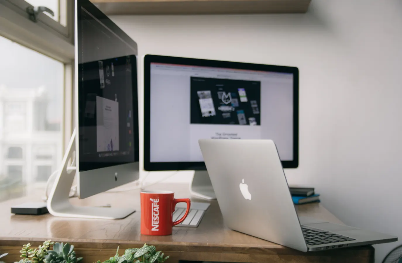 Digital workspace with two desktop monitors, an open laptop, and a red coffee mug