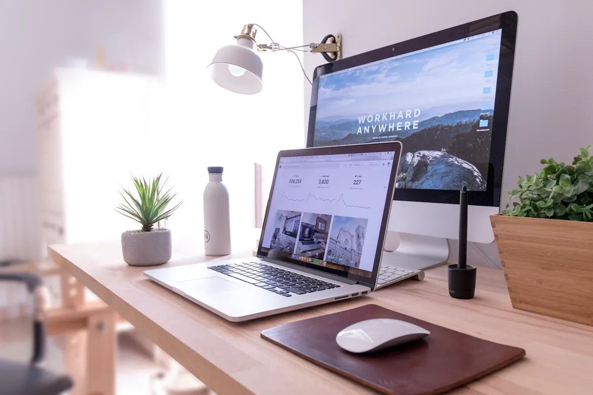 Modern home office workspace with a laptop, desktop monitor, desk lamp, plant, and mouse